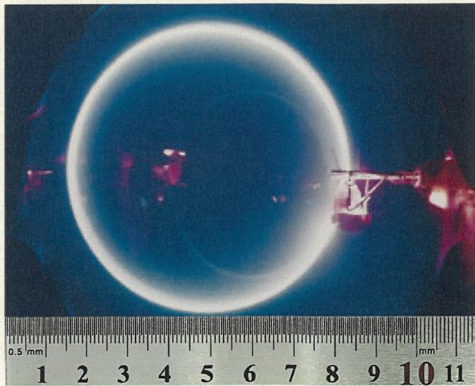 Fotografía de un experimento de rayos catódicos mostrando una trayectoria circular azulada sobre una regla milimetrada para medir el diámetro.